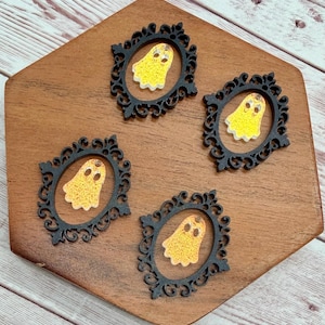 May include: Four black ornate oval frames, each containing a small, glittery yellow ghost figure with two dark eyes. The ghost figures are centered within the open space of the decorative black frames, which feature intricate scrollwork and filigree details.