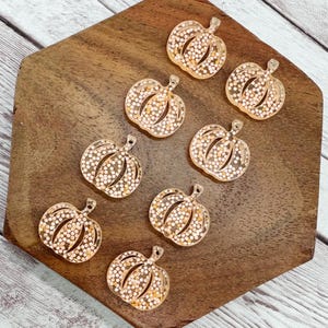 May include: A collection of eight clear acrylic pumpkin charms filled with white and gold glitter. The charms are arranged on a wooden hexagonal tray, perfect for fall-themed crafts or jewelry making.