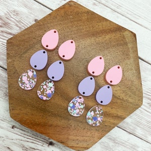 May include: A set of 10 teardrop-shaped resin earrings in pink, purple, and clear with iridescent glitter. The earrings are arranged on a wooden surface.