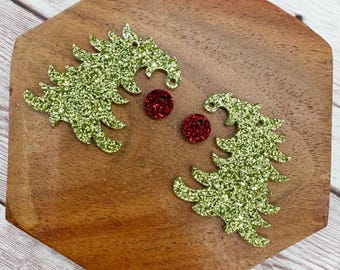 May include: Two glittery green Christmas tree-shaped ornaments and two small red glitter circles are displayed on a wooden surface. The ornaments have a hole at the top for hanging.