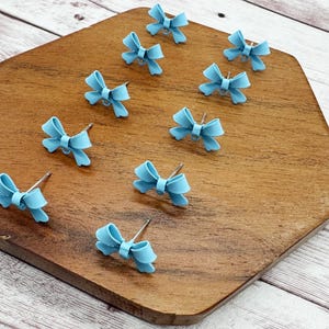 May include: A collection of light blue bow-shaped stud earrings arranged on a wooden surface. The earrings have a simple, elegant design, with a small post for piercing. The wooden surface is a warm brown color, and the background is a light wood.