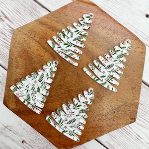 May include: Four Christmas tree-shaped ornaments with a white background and a pattern of green leaves and red berries. Each ornament has a small hole at the top for hanging. The ornaments are on a wooden surface.