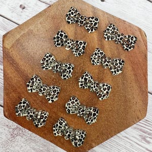 May include: A collection of ten small, bow-shaped hair clips. Each clip features a leopard print pattern in black and brown spots on a cream background. The clips are arranged on a wooden, hexagonal surface.