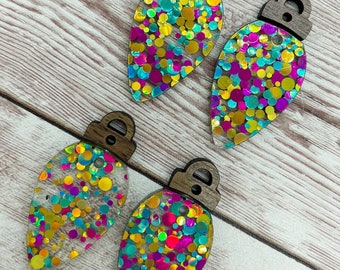 May include: Four clear light bulb ornaments with a wood top. The ornaments are filled with colorful iridescent glitter.