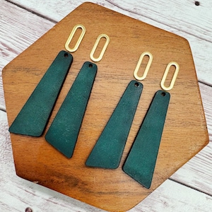 May include: Four dark green leather earrings with gold-colored oval hoops. The earrings are triangular in shape, with a small hole at the top for the hoop. The earrings are displayed on a wooden surface.