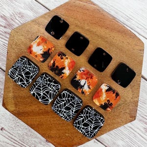 May include: A collection of square Halloween-themed beads. Some are solid black, others feature orange and black tie-dye patterns, and some have a white spiderweb design on a black background. The beads are displayed on a wooden surface.