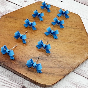 May include: A collection of light blue bow-shaped stud earrings with silver posts. The earrings are arranged on a wooden, hexagonal-shaped surface. The bows have a textured appearance and are evenly spaced.