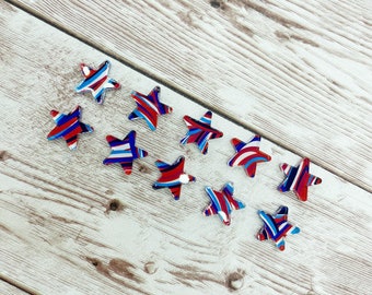Op de afbeelding: Negen stervormige versieringen in rood, wit en blauw met een patroon. De sterren zijn schuin op een licht houten achtergrond gerangschikt.