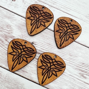 May include: Four brown leather guitar picks with a decorative Celtic knot design. The picks have a hole at the top for hanging.