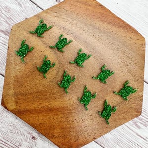May include: A collection of eleven glittery green frog-shaped embellishments arranged on a wooden, hexagonal surface. The frogs are detailed with outstretched limbs, and the wood has a natural grain.