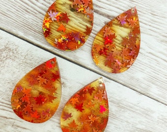 May include: Four teardrop-shaped resin earrings with a wood grain pattern and holographic fall leaves embedded inside. The earrings are a light amber color with red and gold glitter.