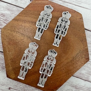 May include: Four silver-colored nutcracker ornaments are arranged on a wooden surface. Each ornament is a detailed cutout of a nutcracker soldier, complete with a crown and uniform. The ornaments are likely for holiday decorations.