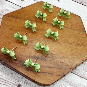 May include: A collection of light green bow-shaped stud earrings displayed on a wooden surface. Each earring features a delicate bow design with a silver post. The earrings are arranged in a scattered pattern.