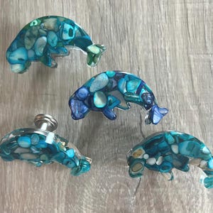May include: Four dolphin-shaped cabinet knobs made of clear resin filled with small, iridescent blue and teal shell fragments. Each knob features a smooth, curved silhouette with a metallic silver-toned mounting base visible on one piece.