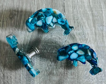 May include: Three dolphin-shaped cabinet knobs featuring a mosaic of blue and white shell fragments embedded in clear resin. Each knob has a smooth, curved surface and a metallic mounting post visible on the side of one of the dolphin pieces.