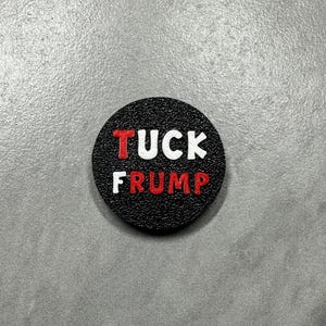 May include: A black, round button with the words "TUCK FRUMP" in red and white lettering. The button has a textured surface and is set against a gray background.