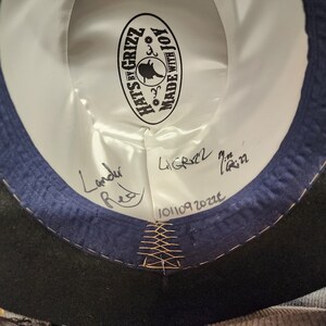 Może przedstawiać: Wnętrze czarnego filcowego kapelusza z niebieską wstążką i białą etykietą z napisem "Hats by Grizz Made with Joy". Etykieta zawiera małą ilustrację czarodziejskiego kapelusza. Kapelusz jest podpisany "Landu Red" i "LiGrizz 7 1/2 in" z datą "101109 2022".