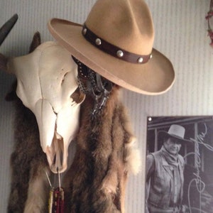 Puede incluir: Un sombrero vaquero de color beige con una banda de cuero marr&oacute;n y tachuelas plateadas. El sombrero est&aacute; colgado en una pared con un cr&aacute;neo y una piel de animal detr&aacute;s. Una foto en blanco y negro de un vaquero es visible en el fondo.