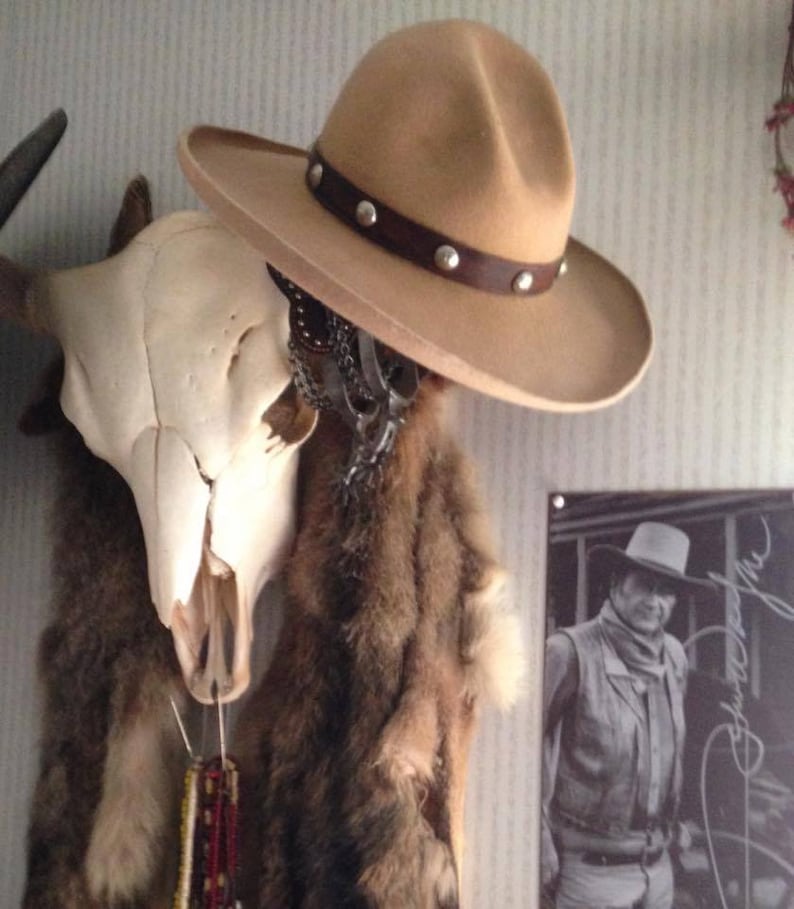 Puede incluir: Un sombrero vaquero de color beige con una banda de cuero marr&oacute;n y tachuelas plateadas. El sombrero est&aacute; colgado en una pared con un cr&aacute;neo y una piel de animal detr&aacute;s. Una foto en blanco y negro de un vaquero es visible en el fondo.