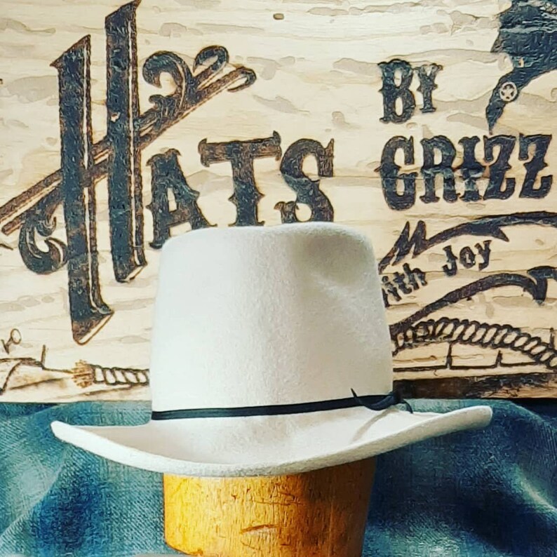 Puede incluir: Un sombrero vaquero de fieltro blanco con una banda negra. El sombrero est&aacute; en un soporte de madera y tiene un ala ancha. El sombrero est&aacute; delante de un letrero de madera que dice "Hats by Grizz with Joy".