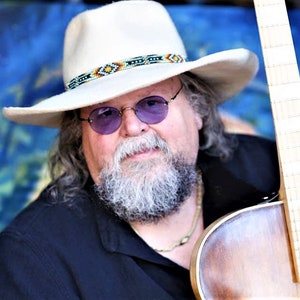 Może przedstawiać: Mężczyzna z długą białą brodą i białym kowbojskim kapeluszem z ozdobnym paskiem z koralik&oacute;w trzyma brązową gitarę akustyczną. Ma na sobie czarną koszulę i złoty naszyjnik.