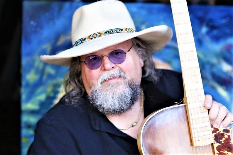 Może przedstawiać: Mężczyzna z długą białą brodą i białym kowbojskim kapeluszem z ozdobnym paskiem z koralik&oacute;w trzyma brązową gitarę akustyczną. Ma na sobie czarną koszulę i złoty naszyjnik.