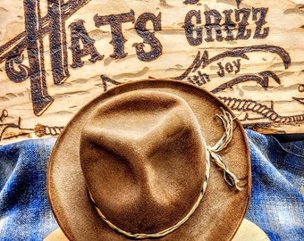 May include: A brown felt fedora hat with a braided leather band. The hat is sitting on a wooden surface with a sign behind it that reads "Hats by Grizz with Joy".