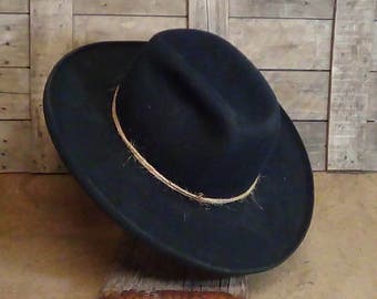 May include: A black felt hat with a wide brim and a pinched crown, featuring a thin, natural-colored twine band wrapped around the base of the crown. The hat has a structured shape and a smooth, matte texture throughout the dark material.
