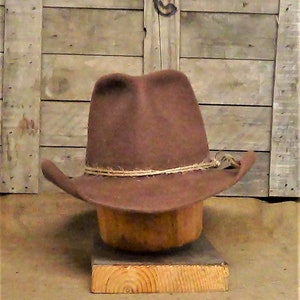Puede incluir: Un sombrero vaquero de fieltro marr&oacute;n con una banda de cuero beige y un acento de cuerda trenzada. El sombrero est&aacute; sentado en un soporte de sombrero de madera.