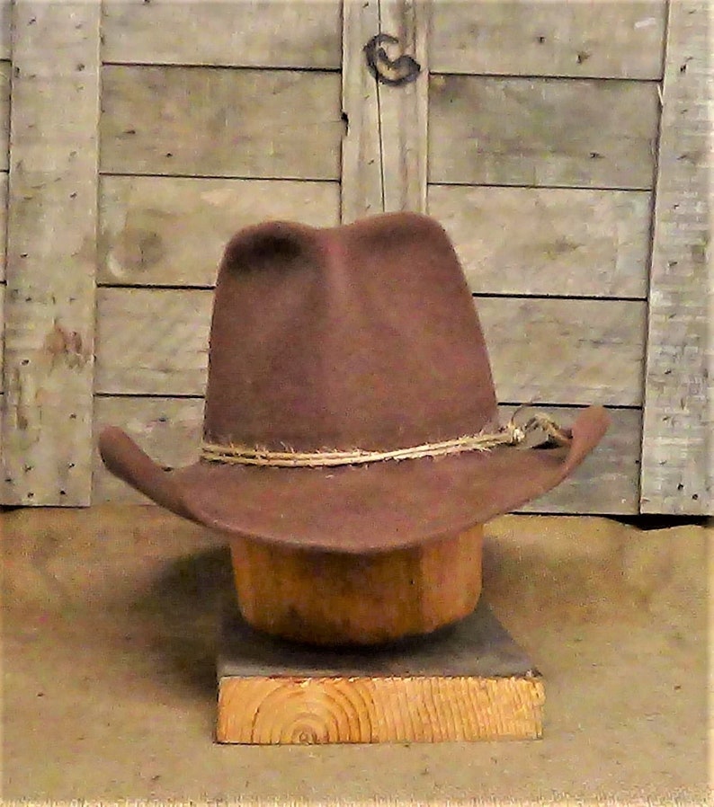 Puede incluir: Un sombrero vaquero de fieltro marr&oacute;n con una banda de cuero beige y un acento de cuerda trenzada. El sombrero est&aacute; sentado en un soporte de sombrero de madera.