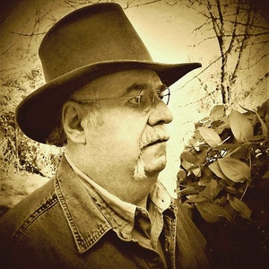 Puede incluir: Un hombre con un sombrero fedora marr&oacute;n y una chaqueta vaquera. Tiene bigote y gafas. El fondo est&aacute; desenfocado y parece ser una zona boscosa.