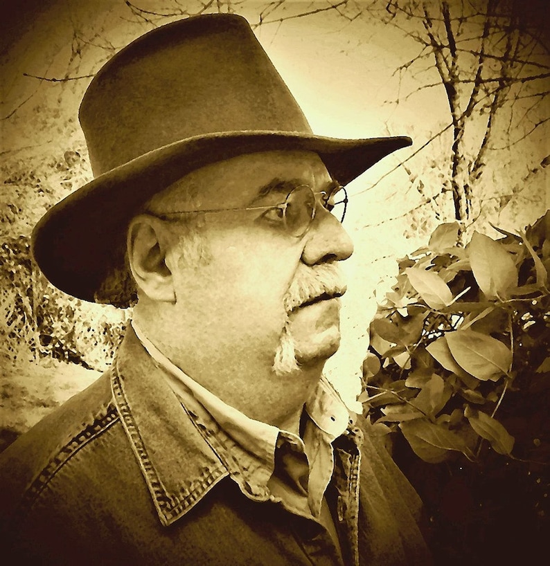 Puede incluir: Un hombre con un sombrero fedora marr&oacute;n y una chaqueta vaquera. Tiene bigote y gafas. El fondo est&aacute; desenfocado y parece ser una zona boscosa.