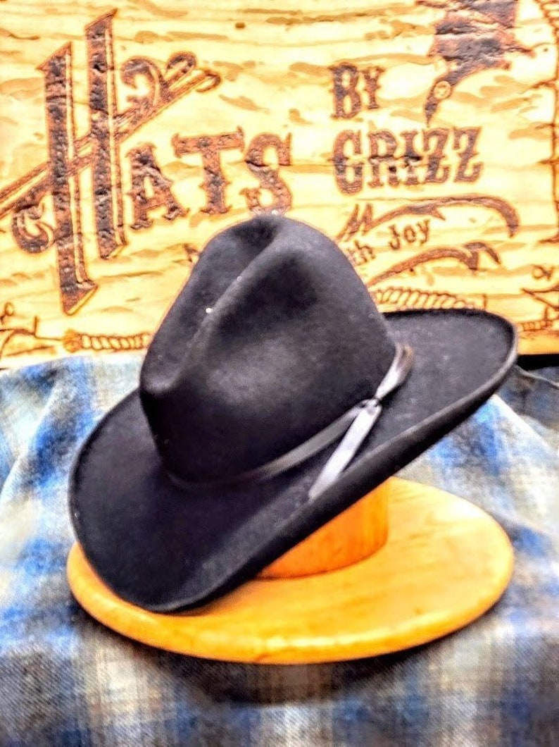 Puede incluir: Un sombrero vaquero de fieltro negro con una banda de cuero marr&oacute;n y una sola pluma. El sombrero est&aacute; sentado en un soporte de madera. El sombrero est&aacute; sobre un fondo a cuadros con un letrero que dice "Hats by Grizz with Joy".