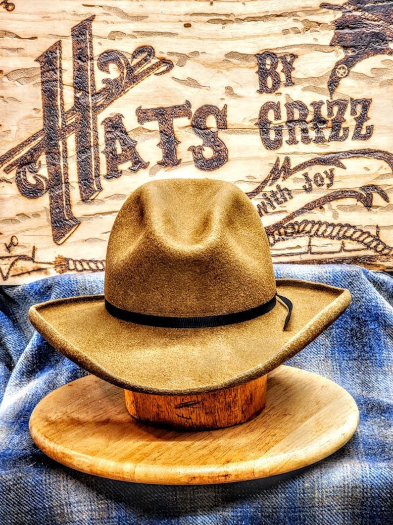 Puede incluir: Un sombrero vaquero de fieltro marr&oacute;n con una banda negra y una cinta de cuero marr&oacute;n. El sombrero est&aacute; sobre un soporte de madera y delante de un letrero de madera que dice "Hats by Grizz with Joy".