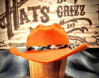 May include: A bright orange cowboy hat with a camouflage band. The hat is on a wooden display stand. The background is a wooden sign with the text "Hats by Grizz".