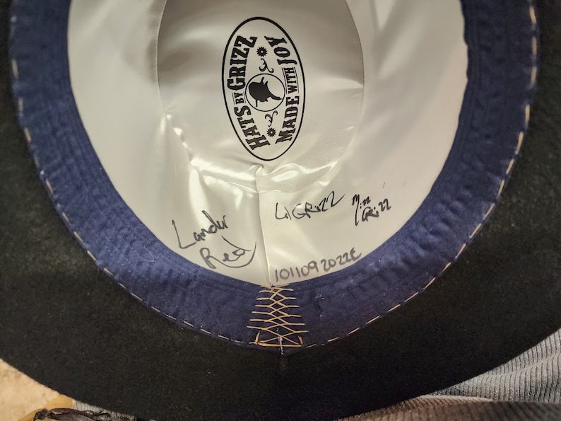 Puede incluir: El interior de un sombrero de fieltro negro con una banda azul y una etiqueta blanca que dice "Hats by Grizz Made with Joy". La etiqueta tiene una peque&ntilde;a ilustraci&oacute;n de un sombrero de bruja. El sombrero est&aacute; firmado "Landu Red" y "LiGrizz 7 1/2 in" con la fecha "101109 2022".