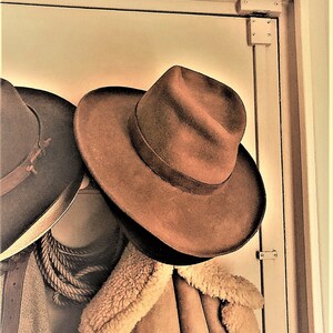 Może przedstawiać: Dwa brązowe filcowe kapelusze kowbojskie wiszące na drewnianej ramie drzwi. Kapelusz po prawej stronie to fedora z szerokim rondem. Kapelusz po lewej stronie to tradycyjny kapelusz kowbojski z węższym rondem.