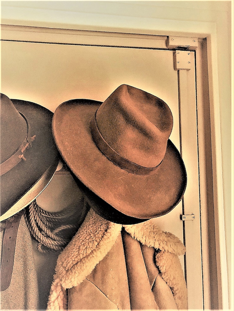 Może przedstawiać: Dwa brązowe filcowe kapelusze kowbojskie wiszące na drewnianej ramie drzwi. Kapelusz po prawej stronie to fedora z szerokim rondem. Kapelusz po lewej stronie to tradycyjny kapelusz kowbojski z węższym rondem.