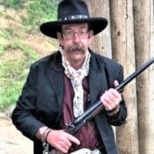 Może przedstawiać: Mężczyzna w czarnym kowbojskim kapeluszu, czarnej kurtce i białym bandanie trzyma karabin z długą lufą. Stoi przed drewnianą konstrukcją.