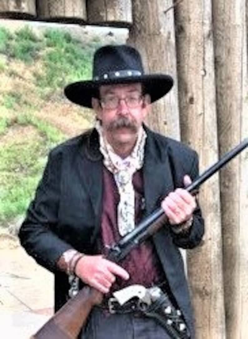 Może przedstawiać: Mężczyzna w czarnym kowbojskim kapeluszu, czarnej kurtce i białym bandanie trzyma karabin z długą lufą. Stoi przed drewnianą konstrukcją.