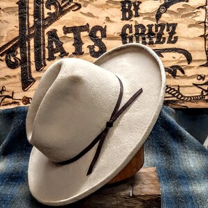 Puede incluir: Un sombrero vaquero de fieltro beige claro con una banda de cuero marr&oacute;n y un lazo. El sombrero est&aacute; sentado en un soporte de madera y un fondo de tela azul a cuadros. El letrero detr&aacute;s del sombrero dice "Hats by Grizz".