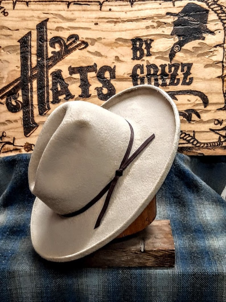Puede incluir: Un sombrero vaquero de fieltro beige claro con una banda de cuero marr&oacute;n y un lazo. El sombrero est&aacute; sentado en un soporte de madera y un fondo de tela azul a cuadros. El letrero detr&aacute;s del sombrero dice "Hats by Grizz".