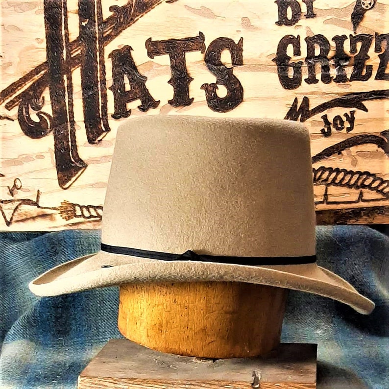 Puede incluir: Un sombrero fedora de fieltro marr&oacute;n claro con una cinta negra. El sombrero est&aacute; sentado en un bloque de sombrero de madera.