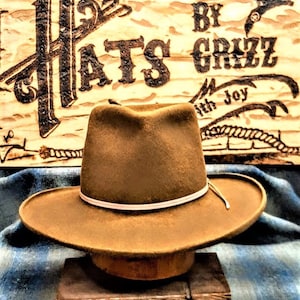 Może przedstawiać: Brązowy filcowy kapelusz kowbojski z białym pasem. Kapelusz stoi na drewnianym bloku przed drewnianym znakiem z napisem "Hats by Grizz with Joy".