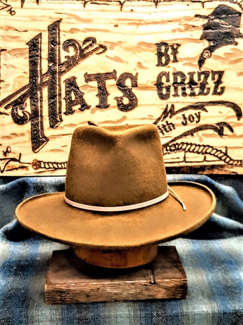 Może przedstawiać: Brązowy filcowy kapelusz kowbojski z białym pasem. Kapelusz stoi na drewnianym bloku przed drewnianym znakiem z napisem "Hats by Grizz with Joy".