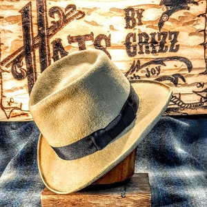 Puede incluir: Un sombrero fedora de color beige con una cinta negra, exhibido en un soporte de madera. El sombrero est&aacute; sobre una tela de mezclilla azul. El fondo es un letrero de madera con el texto "Hats By Grizz" en letras negras.
