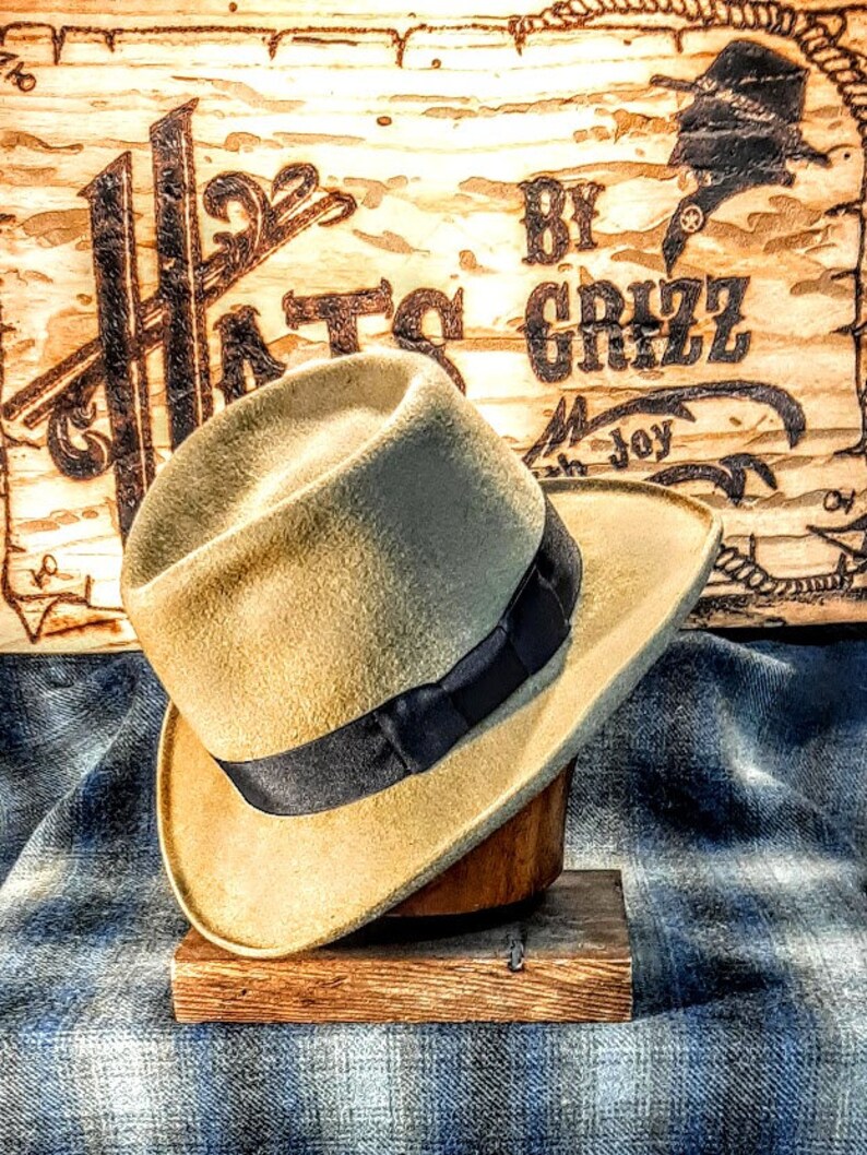 Puede incluir: Un sombrero fedora de color beige con una cinta negra, exhibido en un soporte de madera. El sombrero est&aacute; sobre una tela de mezclilla azul. El fondo es un letrero de madera con el texto "Hats By Grizz" en letras negras.