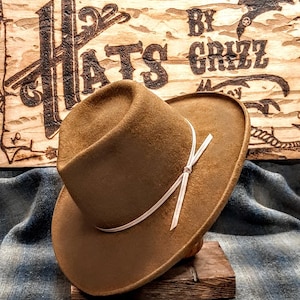 Może przedstawiać: Brązowy filcowy kapelusz fedora z białą wstążką zawiązaną w kokardę. Kapelusz stoi na drewnianym stojaku. Kapelusz jest oznaczony "Hats By Grizz".