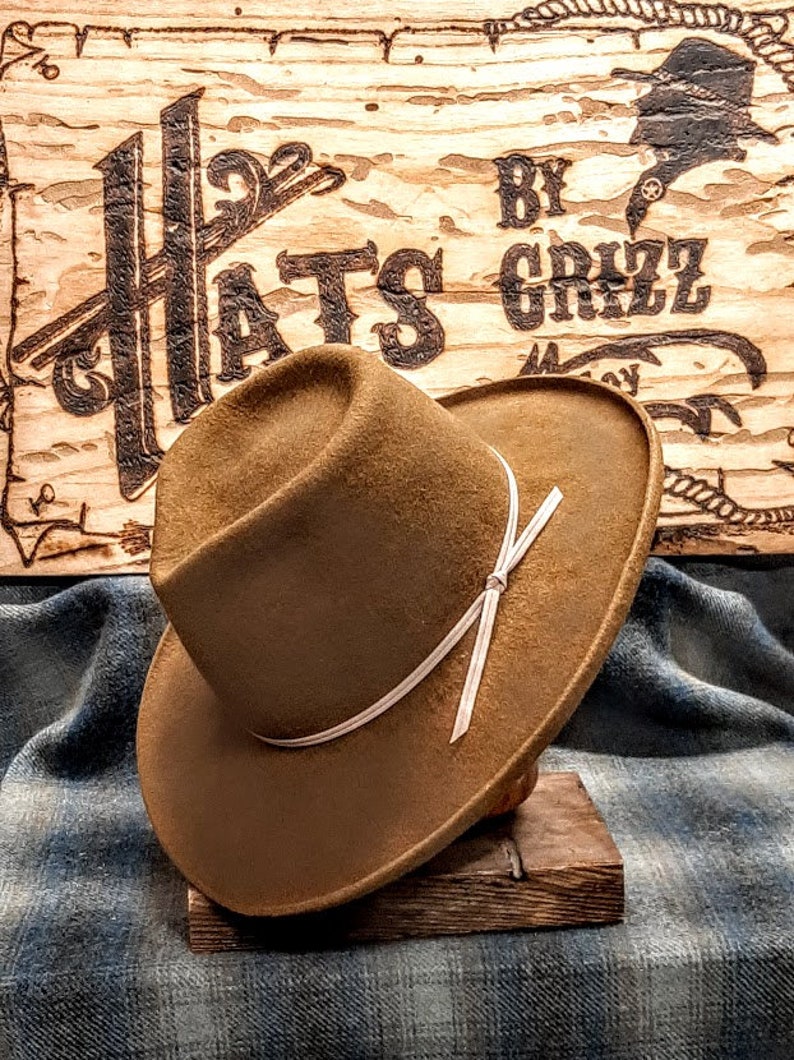Może przedstawiać: Brązowy filcowy kapelusz fedora z białą wstążką zawiązaną w kokardę. Kapelusz stoi na drewnianym stojaku. Kapelusz jest oznaczony "Hats By Grizz".