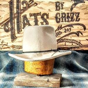 Puede incluir: Un sombrero vaquero de fieltro blanco con una banda negra, exhibido en un soporte de sombrero de madera. El sombrero est&aacute; delante de un letrero de madera con el texto "Hats By Grizz with Joy".